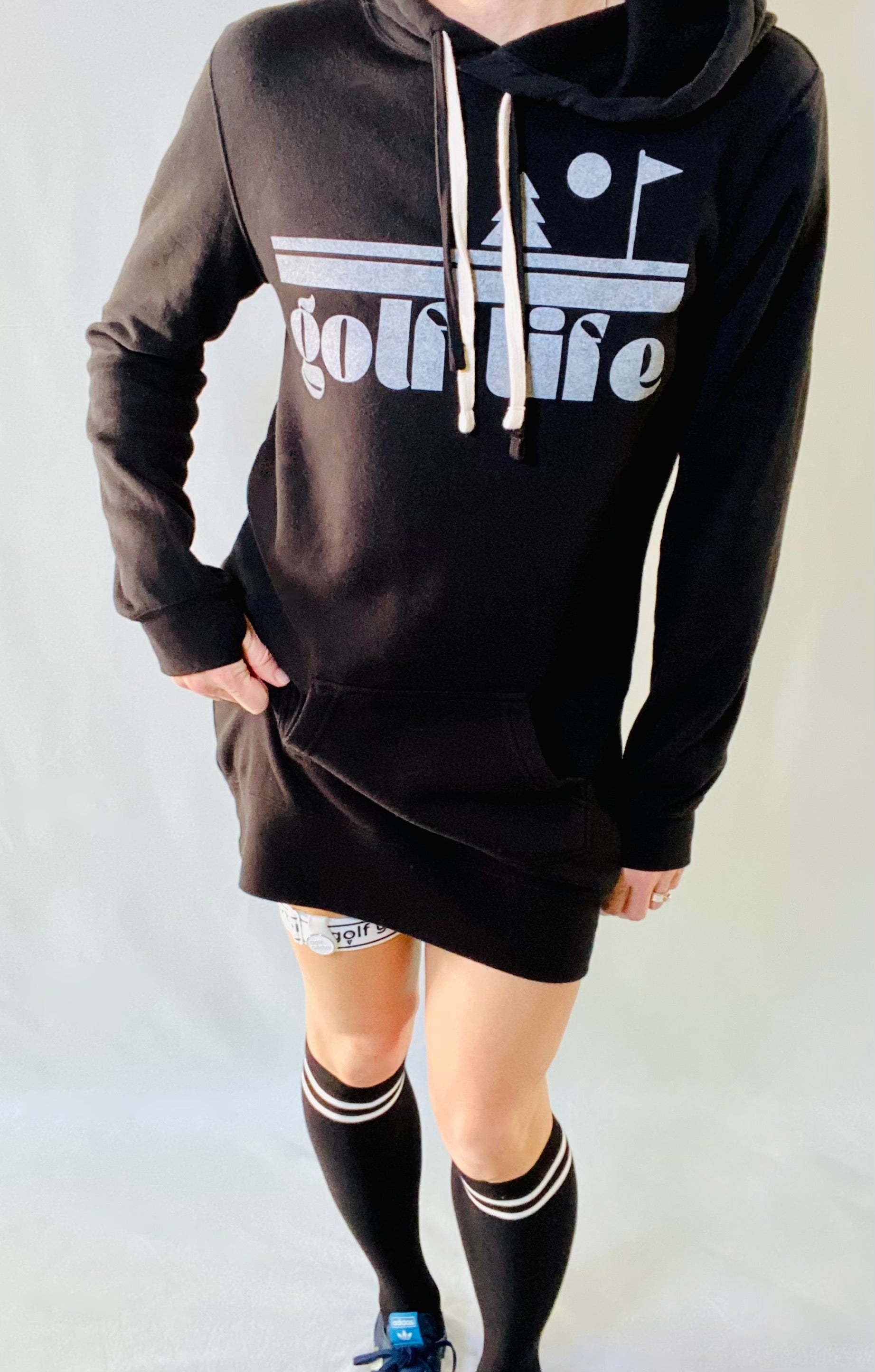 This is a photograph of a woman wearing a black hooded sweatshirt dress. She is wearing black knee-high socks with white stripes. The sweatshirt features a bold design that says golf life. She's lifting her dress a tiny bit to reveal a Golf Garter on her thigh.