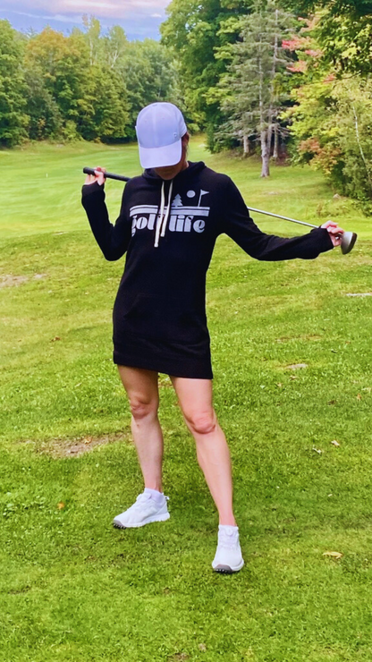 This is a photograph of a woman on the fairway of a golf course. She's wearing a black hooded sweatshirt dress and holding a golf club. She is wearing a white cap and looking down. The sweatshirt dress features a bold design that says golf life.
