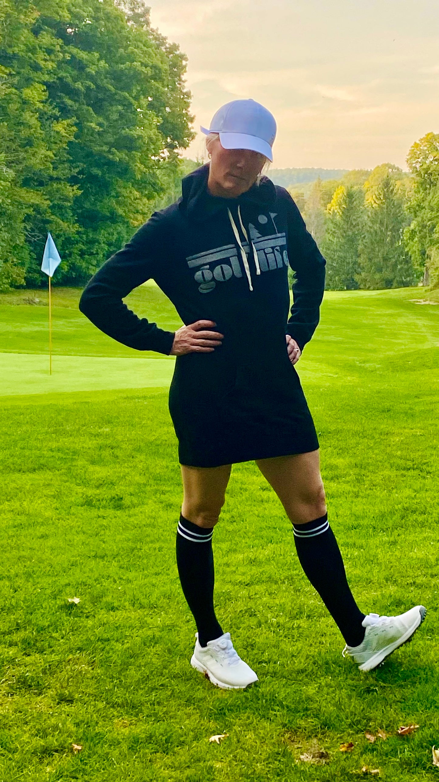 This is a photograph of a woman on a golf course. She is standing near a green with a flag. She's wearing a black hooded sweatshirt dress and knee-high socks. The sweatshirt-dress says golf life.