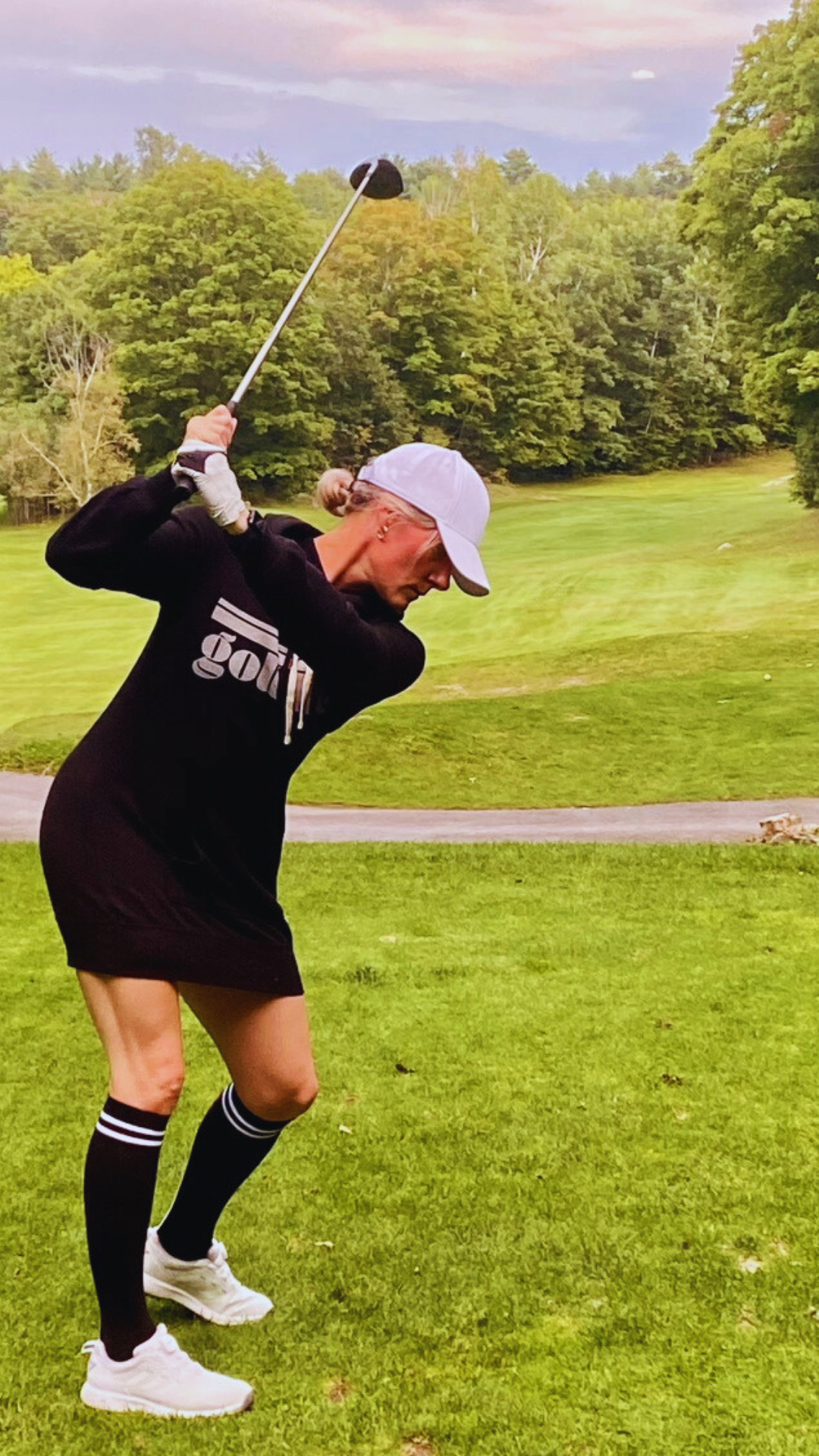 This is a photograph of a woman taking a golf swing on the teebox. She's wearing a black hooded sweatshirt dress, Black knee-high socks and a white baseball hat. The sweatshirt featuring a bold design that says golf life.