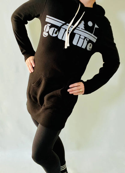 This is a photograph of a woman wearing a black hooded sweatshirt dress over a pair of black leggings. The sweatshirt features a bold design that says golf life.