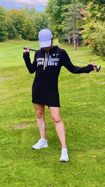 This is a photograph of a woman on the fairway of a golf course. She's wearing a black hooded sweatshirt dress and holding a golf club. She is wearing a white cap and looking down. The sweatshirt dress features a bold design that says golf life.