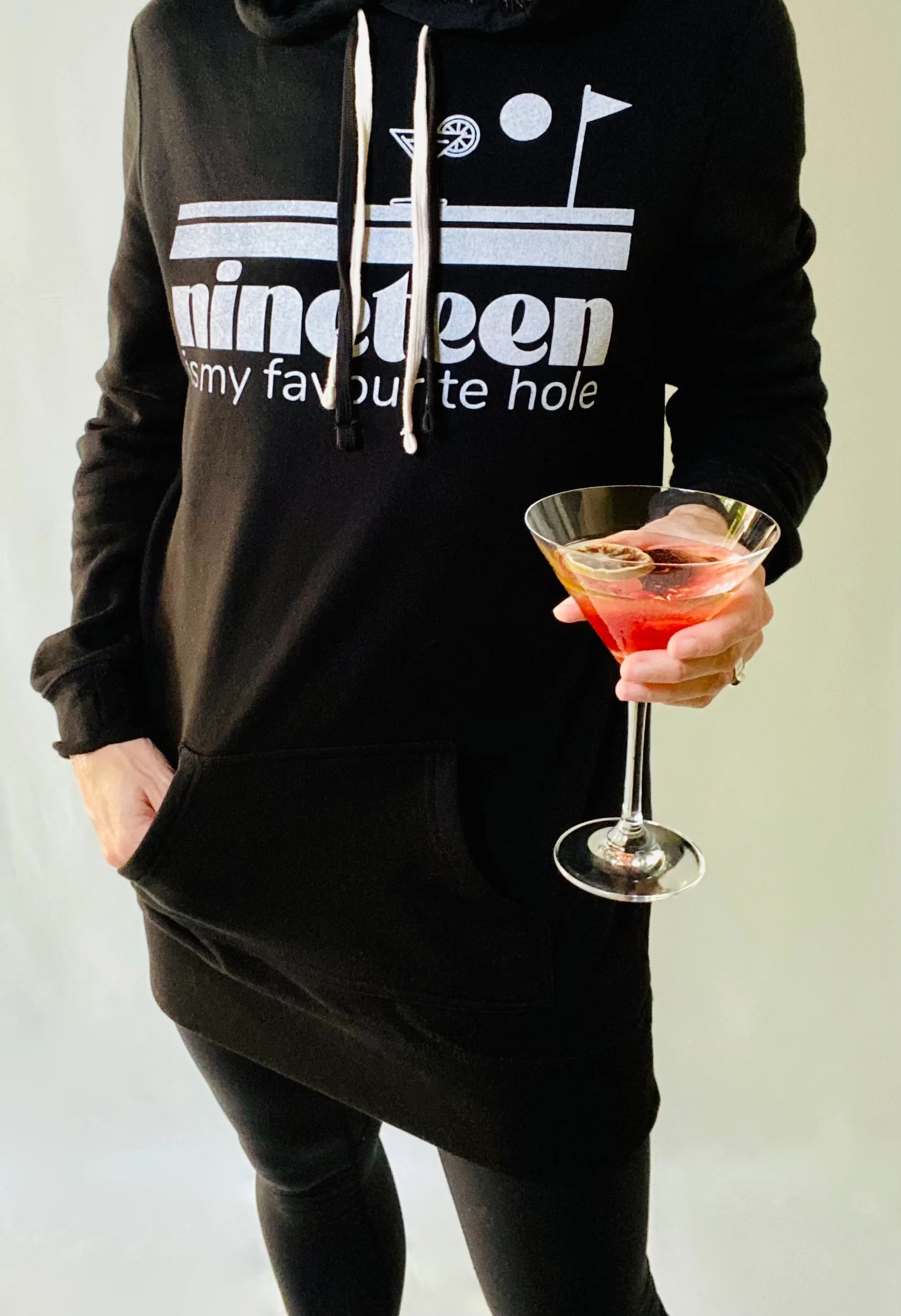 A woman wearing a Nineteenth hole sweatshirt dress by Golf Garter holding a cocktail.