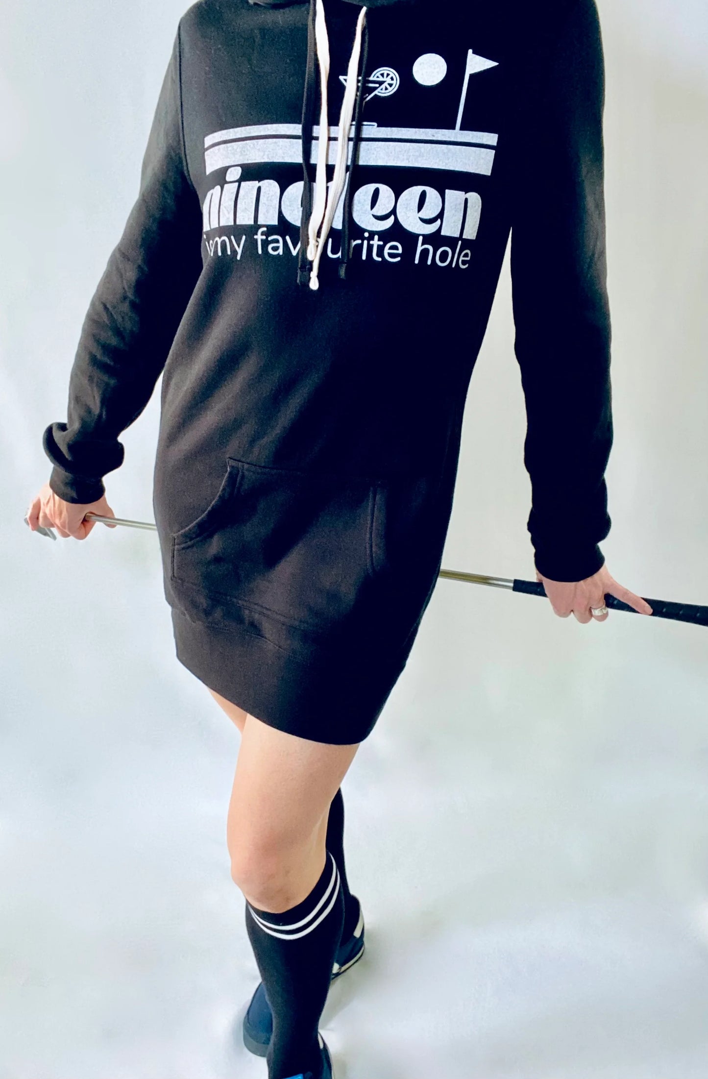 A woman wearing a Nineteenth hole sweatshirt dress by Golf Garter holding a golf club.